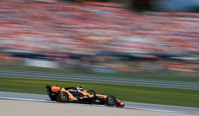 Orange Formula 1 car racing along a track with a crowded, blurred grandstand in the background (motion blur).