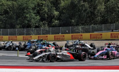 A dense pack of Formula 1 cars racing on the track, with Pirelli banners along the fence in the background on a sunny day.