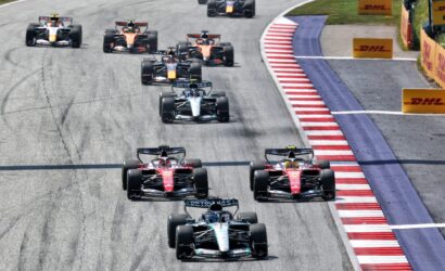 Aerial view of a Formula One race with a pack of cars navigating a curved track, DHL banners along the barriers on the right.