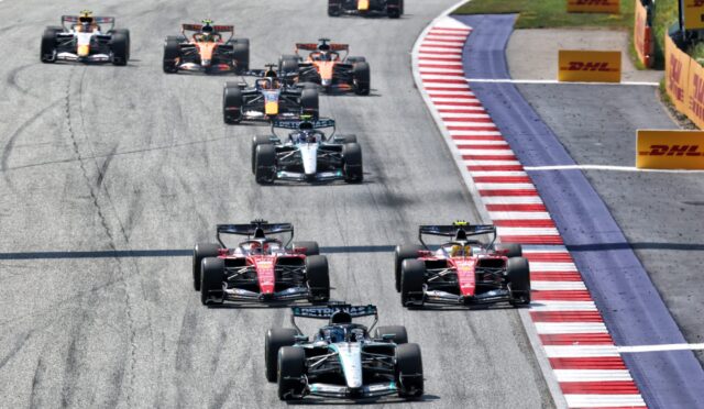Aerial view of a Formula One race with a pack of cars navigating a curved track, DHL banners along the barriers on the right.