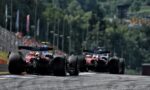 Three Formula 1 cars seen from behind speeding down a race track toward the camera with a blurred, forested backdrop behind them.