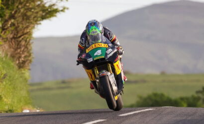 MotoGP rider in full racing gear airborne over a rural road with hills in the background.
