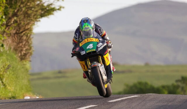 MotoGP rider in full racing gear airborne over a rural road with hills in the background.