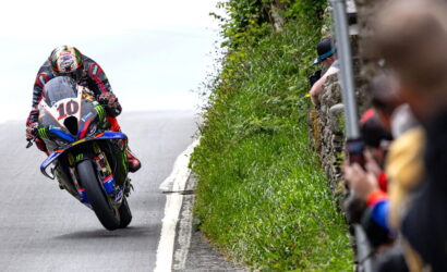 Motorcycle racer leaning into a left-hand turn on a track, bike #10, with spectators behind a stone wall on the right side.