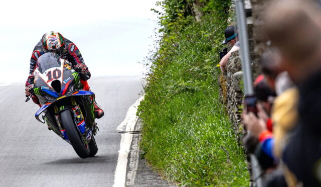 Motorcycle racer leaning into a left-hand turn on a track, bike #10, with spectators behind a stone wall on the right side.