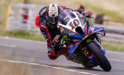 MotoGP rider in racing suit leans into a curve on a blue/black bike number 10, speed blurred asphalt track in background, sponsor logos visible.