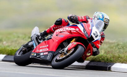 Motorcycle racer in red livery leans into a curve on a racetrack, number 3, with sponsor logos on the bike.