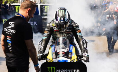 Moto racer in full leathers and helmet on a blue-yellow race bike, crew member in a SCARS Racing shirt watching as smoke circles them at the track edge.