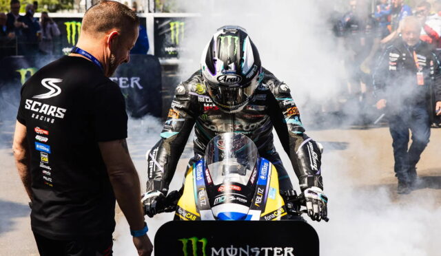 Moto racer in full leathers and helmet on a blue-yellow race bike, crew member in a SCARS Racing shirt watching as smoke circles them at the track edge.