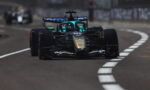 Front-facing Formula 1 car in black and turquoise, racing on a curved track with white dashed lines.