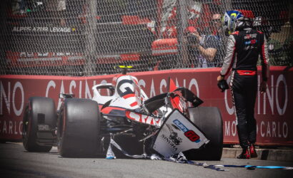 Monaco race crash scene: a damaged open-wheel car sits on the track with debris scattered, a crew member in a racing suit stands nearby behind a safety barrier and photographers observe from behind the fence.