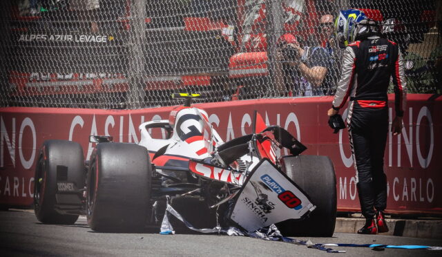Monaco race crash scene: a damaged open-wheel car sits on the track with debris scattered, a crew member in a racing suit stands nearby behind a safety barrier and photographers observe from behind the fence.