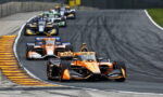 Orange open-wheel race car leads a pack around a curve on a race track, with tire marks visible behind the cars.