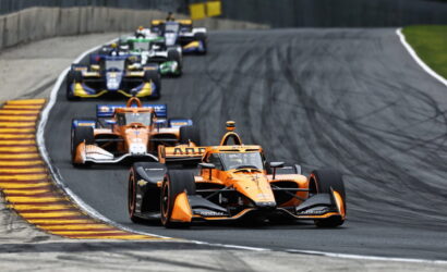 Orange open-wheel race car leads a pack around a curve on a race track, with tire marks visible behind the cars.