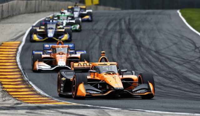 Orange open-wheel race car leads a pack around a curve on a race track, with tire marks visible behind the cars.