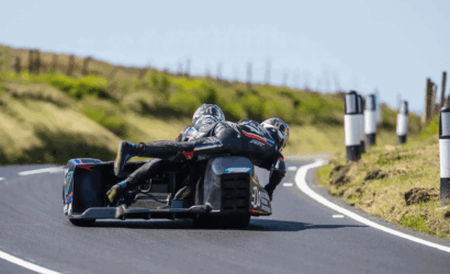 Two riders lean into a sharp turn on a sidecar race bike on a winding rural road