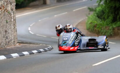 Two riders on a blue and red sidecar racing motorcycle lean into a sharp turn on a winding road.