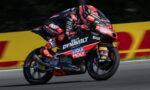 Rider in red‑black race suit leaning into a curve on a sponsor‑liveried racing motorcycle on a track, with DynamoLt and Liqui Moly logos visible.