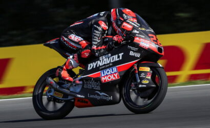 Rider in red‑black race suit leaning into a curve on a sponsor‑liveried racing motorcycle on a track, with DynamoLt and Liqui Moly logos visible.