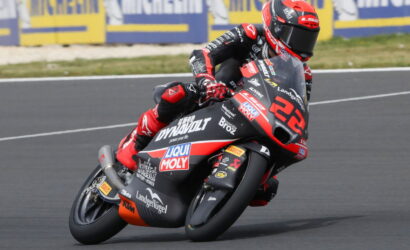 Racer on a red-and-black motorcycle leans into a sharp turn on a racetrack, sponsor logos visible on the bike and suit