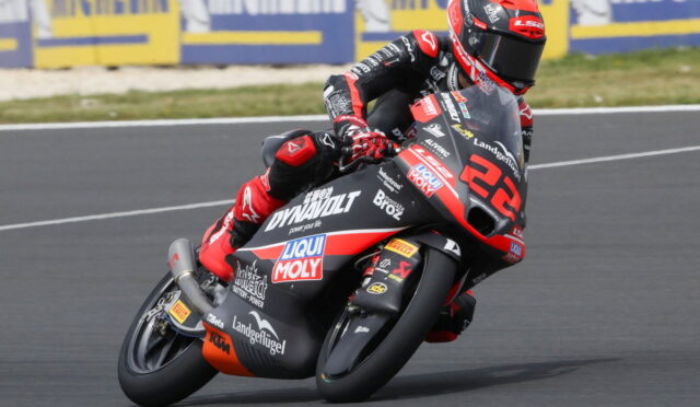 Racer on a red-and-black motorcycle leans into a sharp turn on a racetrack, sponsor logos visible on the bike and suit