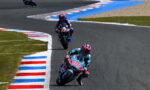 Motorcycle racers on a curving track, rider #80 in teal leading, followed by rider #17 on a blue bike, with red‑white‑blue curbs lining the corner.