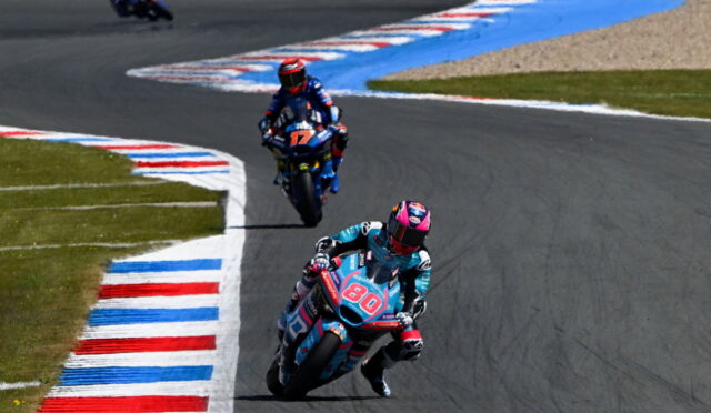 Motorcycle racers on a curving track, rider #80 in teal leading, followed by rider #17 on a blue bike, with red‑white‑blue curbs lining the corner.