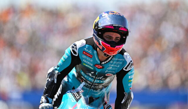 Motorcycle racer in turquoise suit and pink helmet leaning into a turn on a blue bike during a race, with a blurred crowd in the background.