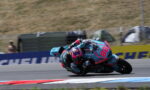 Motorcycle racer in teal and pink gear leans into a sharp turn on a race track, bike numbered 84.