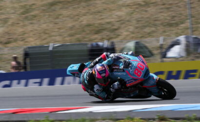 Motorcycle racer in teal and pink gear leans into a sharp turn on a race track, bike numbered 84.