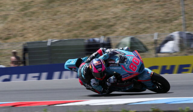 Motorcycle racer in teal and pink gear leans into a sharp turn on a race track, bike numbered 84.