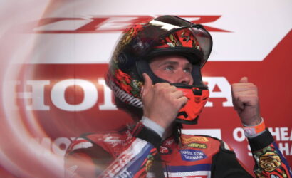 Racer in red racing suit wearing a fiery helmet, giving a thumbs-up on a red sponsor backdrop.
