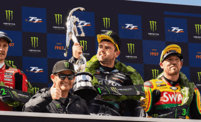 Three motorcycle racers on a podium with a large trophy, set against a Monster Energy backdrop and laurel garlands.