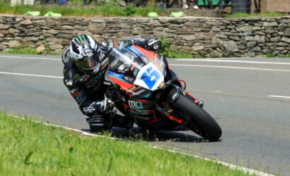 Motorcycle racer in a black racing suit leans into a turn on a public road with a stone wall and grass in the background.