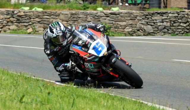 Motorcycle racer in a black racing suit leans into a turn on a public road with a stone wall and grass in the background.