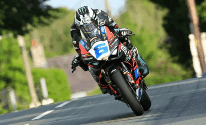 Motorcycle racer in full gear leans into a curve on a sports bike with number plate on a rural road.