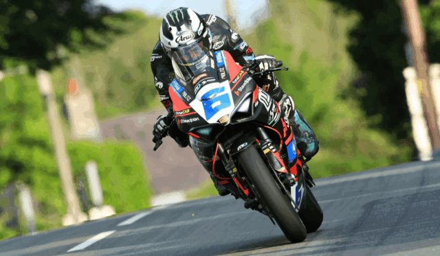 Motorcycle racer in full gear leans into a curve on a sports bike with number plate on a rural road.