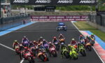 Group of motorbike racers leaning into a start on a race track with MotoGP banners overhead in the stadium area.