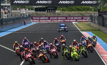 Group of motorbike racers leaning into a start on a race track with MotoGP banners overhead in the stadium area.