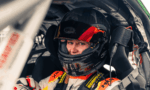 Racing driver in a Bell helmet with red visor sits in a race car cockpit, looking at the camera with harness fastened.