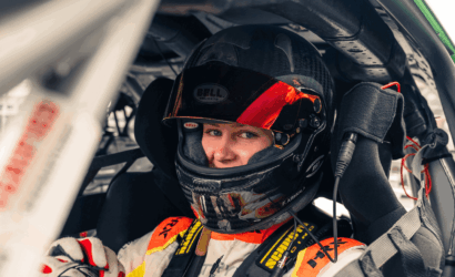 Racing driver in a Bell helmet with red visor sits in a race car cockpit, looking at the camera with harness fastened.