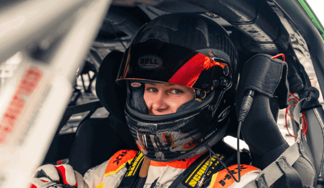 Racing driver in a Bell helmet with red visor sits in a race car cockpit, looking at the camera with harness fastened.