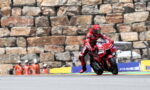 Red-suited motorcycle racer leans into a turn on a track, with a stone wall and orange-clad officials behind.