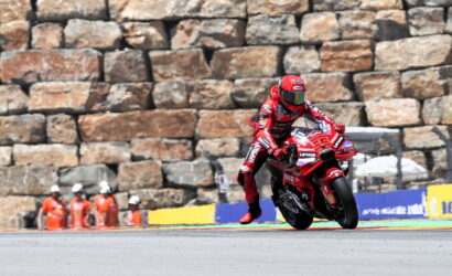 Red-suited motorcycle racer leans into a turn on a track, with a stone wall and orange-clad officials behind.