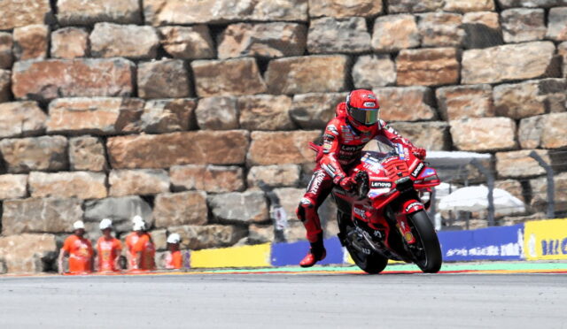 Red-suited motorcycle racer leans into a turn on a track, with a stone wall and orange-clad officials behind.