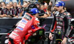 Two MotoGP riders in racing gear stand beside a red racing bike as a crowd and cameras surround them in a pit area scene.