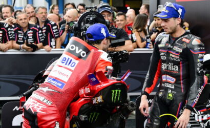 Two MotoGP riders in racing gear stand beside a red racing bike as a crowd and cameras surround them in a pit area scene.