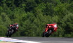 Two motorcycle racers in red and black leathers lean into a sharp turn on a race track with a dense green forest in the background.