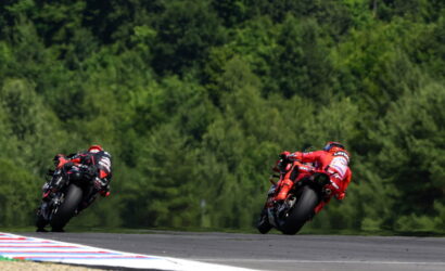Two motorcycle racers in red and black leathers lean into a sharp turn on a race track with a dense green forest in the background.