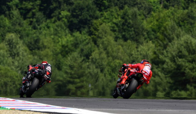 Two motorcycle racers in red and black leathers lean into a sharp turn on a race track with a dense green forest in the background.